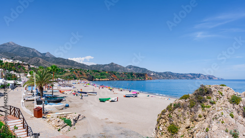 Playa con una gran roca y paseo marítimo ,con barcas sobre la arena, el mar, acantilados y montañas de fondo un día soleado de vacaciones en verano, con cielo azul. Desde Málaga, Andalucía, España. photo