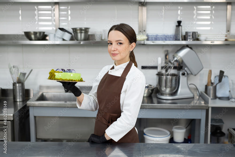 Young smiling brunette chef holds a mousse cake decorated with handmade chocolates.