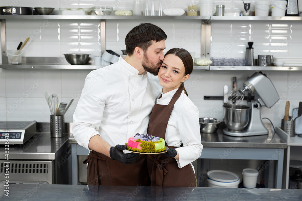 Happy loving couple of pastry chefs, a man and a woman hold a mousse cake in their hands in a professional kitchen. Happy loving couple of pastry chefs, a man and a woman hold a mousse cake in their hands in a professional kitchen.