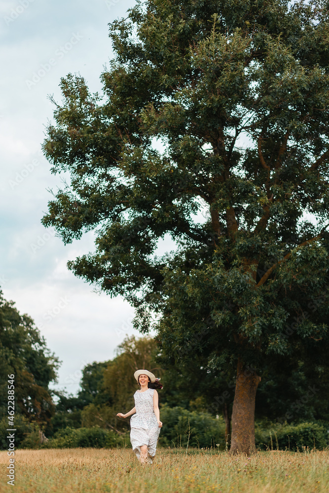 A happy young woman in a dress and a straw hat runs across the field. In the background is a large tree and a stormy sky. Vertical