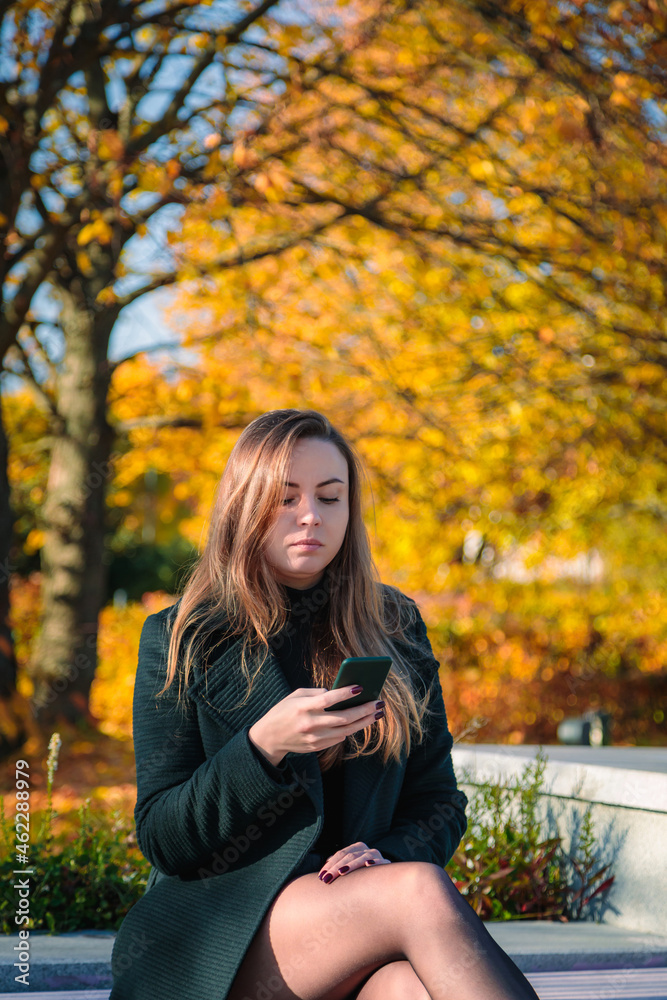 A woman with long hair sits on a bench in an autumn park and looks into a mobile phone. A woman with long hair sits on a bench in an autumn park and looks into a mobile phone.