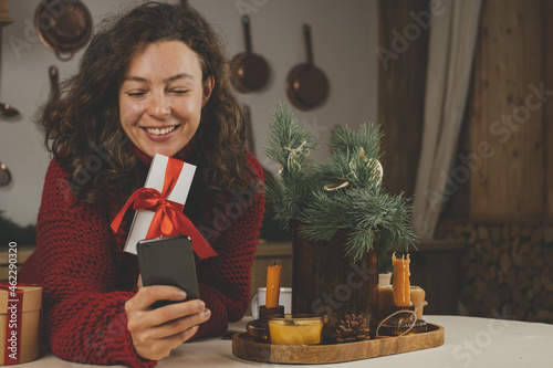 Smiling brunette woman choosing christmas gifts, using smartphone, looking at screen, sitting near festive tree indoor,wearing red sweater. Shopping online, enjoying winter holiday, browsing website photo