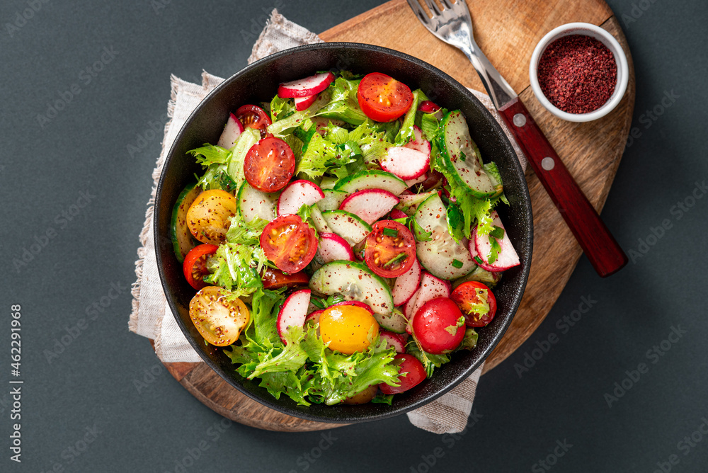 Spring vegetable salad with olive oil and sumac in a black bowl on a dark background. Salad of radishes, cucumbers, tomatoes, lettuce, sumac, and olive oil. Vegan or diet food. Healthy lunch.