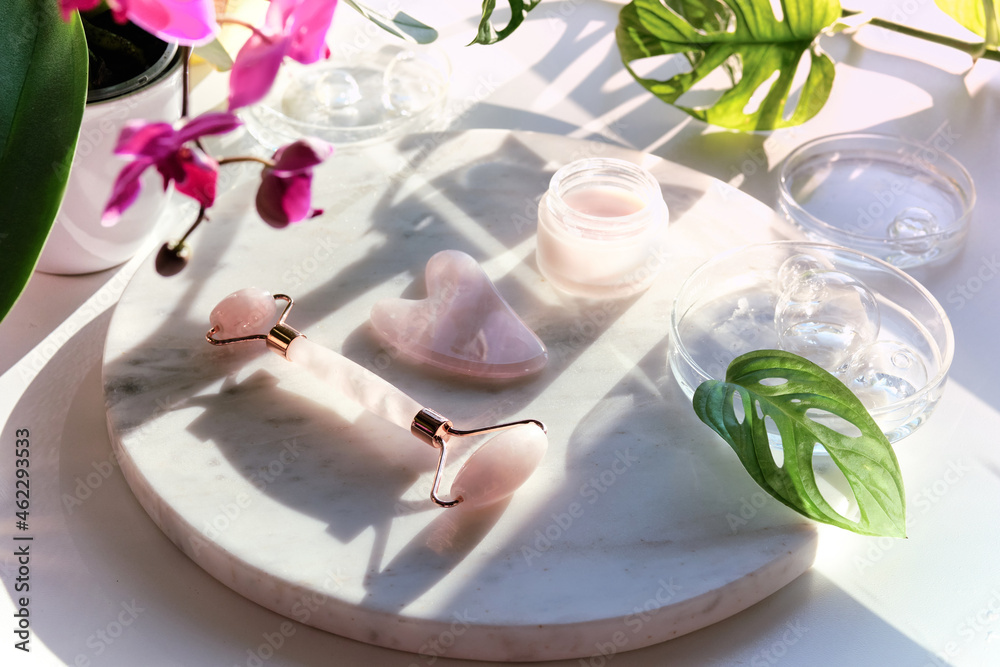 Moisturizer, pink quartz face roller with exotic monstera leaves and orchid flowers. Sunshine, long shadows. Facial massage background with self made cream on marble plate.