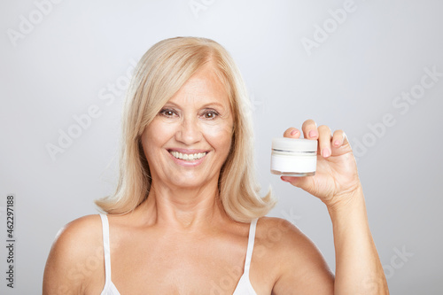 A happy senior blond woman holding anti-age creme in hand and looking at the camera.