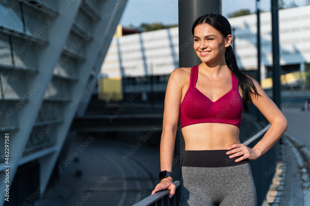 Healthy brunette woman looking away and smiling widely while spending time at the stadium after hard training. Girl standing in front of the camera. Sport concept