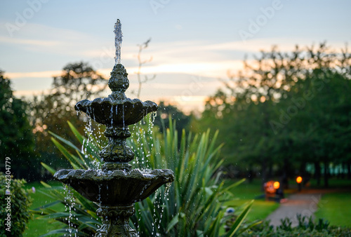 Fountain with water drops in a park with trees behind at dusk. The fountain is seen with a path, bench and lights just after sunset. This fountain is a central feature in the park.