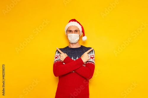 man in Christmas sweater with reindeer, medical mask, red Santa Claus hat points with his index fingers in different directions at an empty place for New Year advertising. isolated yellow background