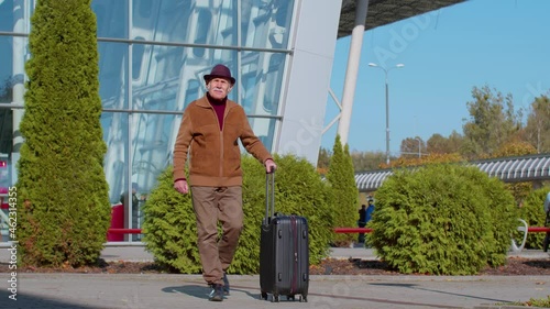 Lovely stylish mature retired grandfather businessman walking with luggage suitcase bag to airport hall or railway station for boarding. Travel, vacation, journey, trip, holiday. Senior pensioner man