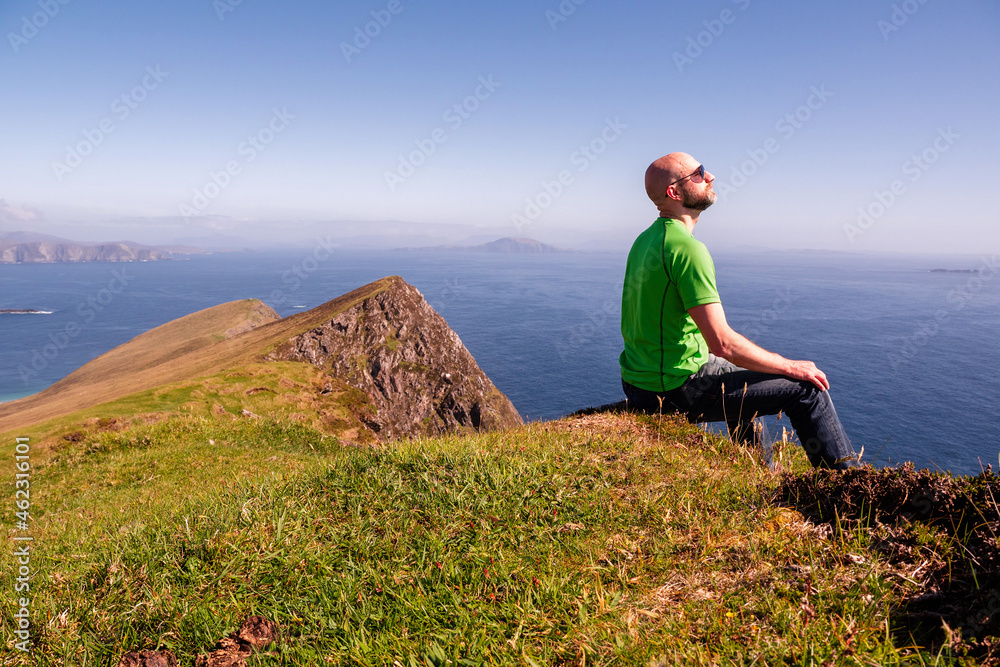 Bald man relaxing and enjoys view on the ocean from a mountain peak, warm sunny day. Male tourist, slim body, wearing green t shirt and blue jeans. Achill island, county Mayo, Ireland.