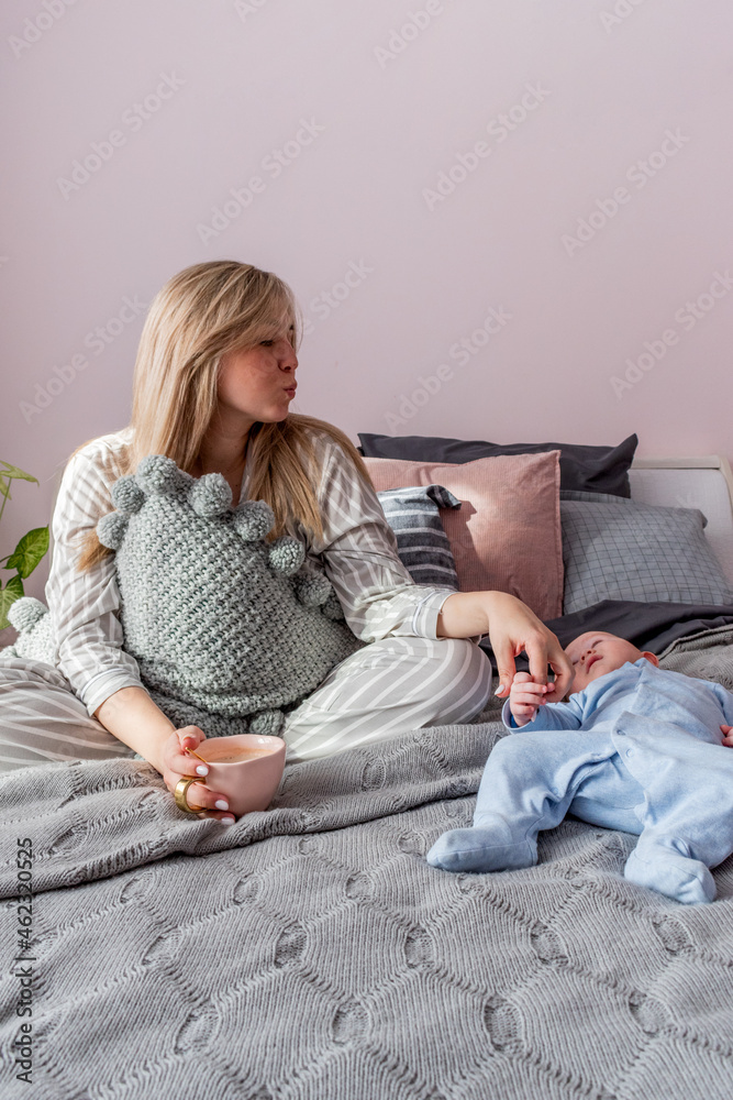 Obraz premium Young woman sits on bed in bedroom and drinks coffee next to her is infant.