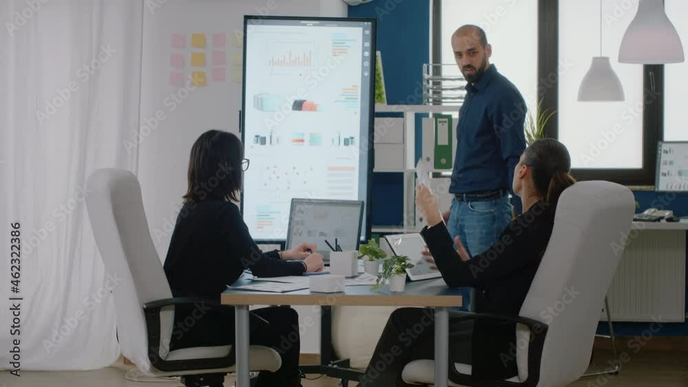 Man pointing at television with chart presentation for project planning while businesswomen listening to ideas for marketing strategy at meeting. Workmates with research development Man pointing at television with chart presentation for project planning while businesswomen listening to ideas for marketing strategy at meeting. Workmates with research development