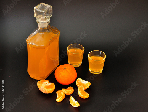 Homemade tangerine liqueur, a glass bottle and two glasses with alcohol on a black background, next to the ripe citrus slices.