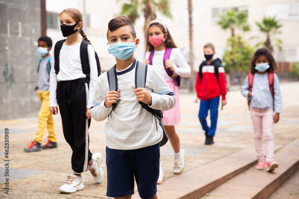Obraz premium Portrait of schoolboy in medical mask standing near school, kids on background