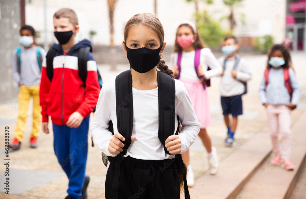 Obraz premium Confident teen girl in protective mask walking outside school building on spring day, going to lessons. Concept of necessary precautions in COVID pandemic.