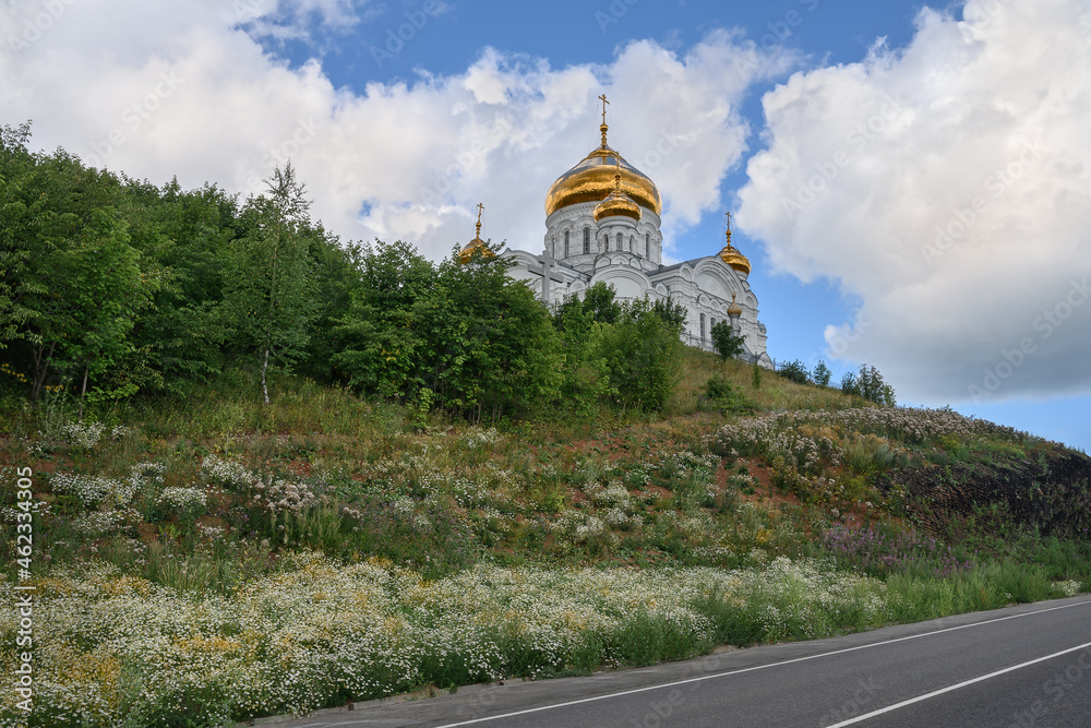The majestic white church with golden domes on White Mountain (Perm Territory, Russia) on a cloudy summer day. The church is surrounded by beautiful nature and wildflowers. The road to the temple 