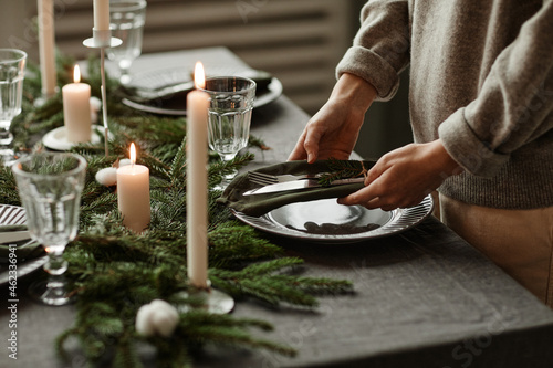 Side view close up of unrecognizable woman setting up dining table decorated for Christmas with fir branches and candles in grey tones, copy space