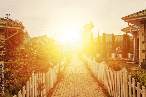 Sidewalk between cottages and gardens to rising sun. Small fences around road. Nice houses and trees. Summer landscape. Bright sunlight at end of street.