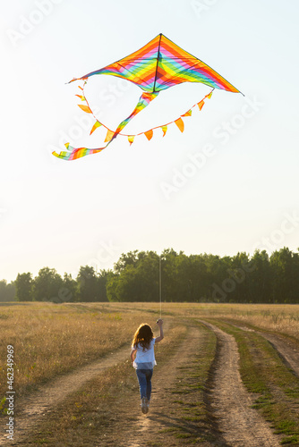 Little girl with rainbow colored kite running along the country road in the summer field. Selective focus.
