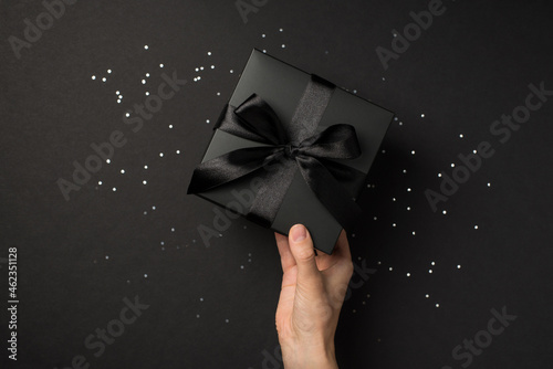 First person top view photo of hand holding stylish black giftbox with black ribbon bow over shiny sequins on isolated black background