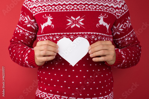Cropped closeup photo of young man in red and white christmas sweater holding white paper heart on isolated red background with blank space © ActionGP