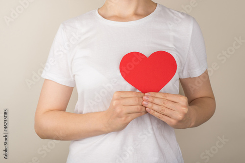 Fototapeta Naklejka Na Ścianę i Meble -  Cropped closeup photo of young woman in white t-shirt holding red paper heart near heart on isolated grey background