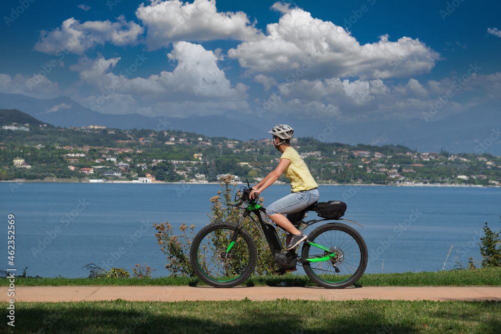 The movement of an electric cyclist along the coastline in the background lake, mountains, blue sky with clouds. Woman on a sports mountain electric bike lake garda.