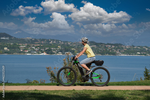 The movement of an electric cyclist along the coastline in the background lake, mountains, blue sky with clouds. Woman on a sports mountain electric bike lake garda.