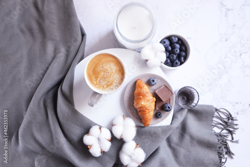 Cup of coffee with croissant, chocolate bar, blueberries, cotton flowers and grey scarf on the white marble background. Autumn, winter coffee break. Cozy warm home latte. Relax at home.