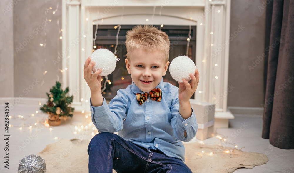 Naklejka premium A Caucasian little boy in a blue shirt and a tiger bow tie is sitting near the fireplace in a room with Christmas balls