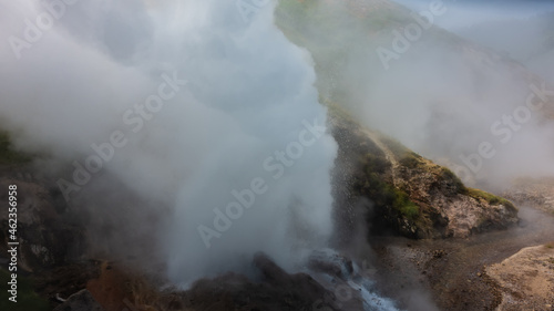A column of thick steam rises from the cauldron of an erupting geyser, splashes of boiling water scatter around. The hillside is shrouded in fog. Kamchatka. Valley of geysers.