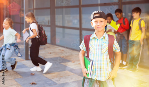 Cute cheerful tween boy looking at camera while standing in school campus with notebooks in hands on background with schoolmates.