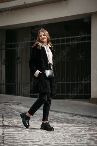 Close-up photo of young beautiful woman in black fur coat, jeanse and shoes posing on street photo