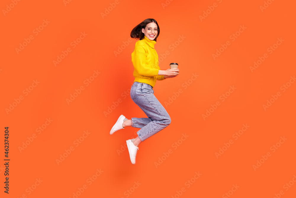 Full length profile side photo of young woman happy positive smile jump takeaway coffee isolated over orange color background Full length profile side photo of young woman happy positive smile jump takeaway coffee isolated over orange color background