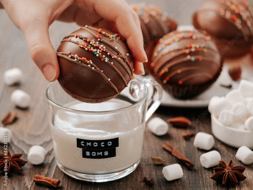 Chocolate cocoa bomb in hand near glass cup with plant-based milk and choco bomb text. Ball made from milk chocolate with marshmallow. Stylish orange toned image of trendy winter hot chocolate drink - obrazy, fototapety, plakaty 