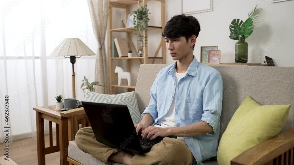 asian male freelance worker sitting cross legged on sofa while working on the computer with concentration in a bright living room at home