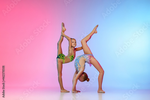 Portrait of two little beautiful rhythmic gymnastics artists, girls in sport event isolated on gradient pink blue studio background in neon light