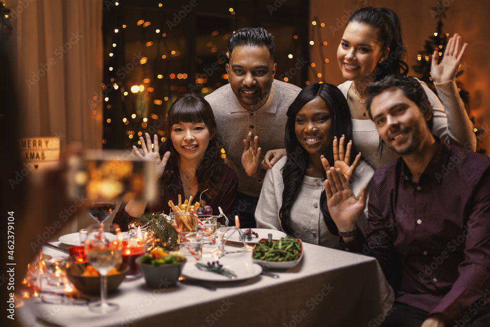 holidays and celebration concept - multiethnic group of happy friends photographing at christmas dinner and waving hand