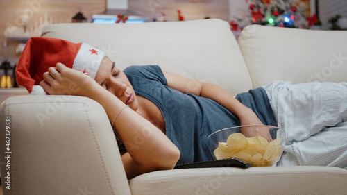 Close up of adult sleeping on couch with bowl of chips and TV remote control. Woman wearing santa hat resting on christmas holiday at television screen. Person alseep on sofa. photo