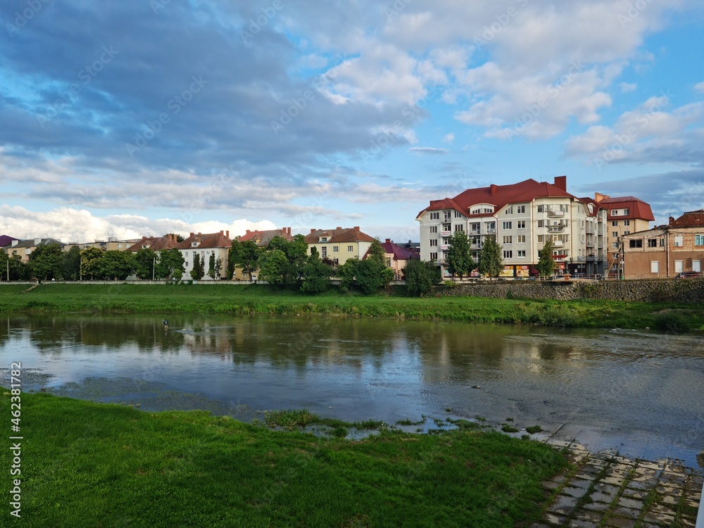 Natural beauty of blue sky and calm water of river in Mukachevo, Ukraine. Beautiful landscape with river and houses at sunset. Scenic view of the old town embankment on a cloudy summer day at dusk. Natural beauty of blue sky and calm water of river in Mukachevo, Ukraine. Beautiful landscape with river and houses at sunset. Scenic view of the old town embankment on a cloudy summer day at dusk.