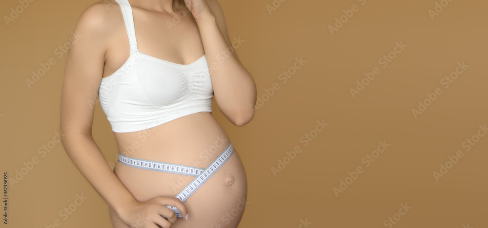Beautiful pregnant young woman in clothes for pregnant women is measuring her bare tummy on a biege background. Pregnant woman measuring her large belly