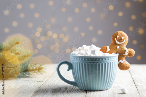 A blue cup of winter hot drink with marshmallows and gingerbread man on a white wooden background with bokeh.
