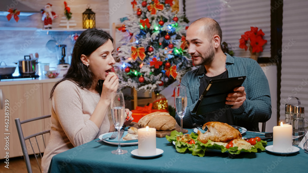 Festive couple looking at tablet while enjoying christmas dinner, celebrating and clinking glasses of champagne. Man and woman using device, enjoying traditional holiday meal and alcohol