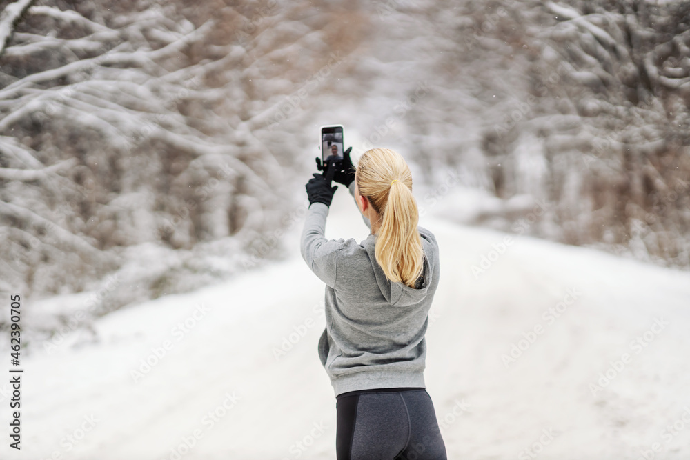 Rear view of sportswoman standing in nature at snowy winter day and taking a selfie. Technology, trends, winter fitness