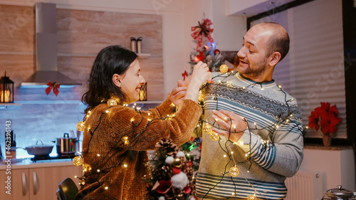 Festive couple trying to untangle garland of twinkle lights while decorating kitchen. Man and woman feeling cheerful and getting tangled in string of christmas lights with illuminated bulbs photo