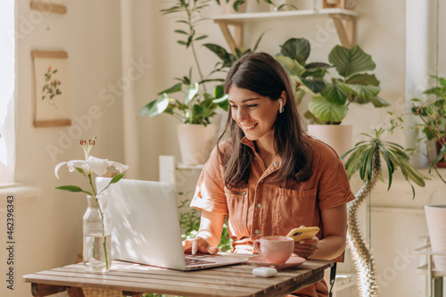 Obraz na płótnie Positive young mixed race woman using a laptop and smartphone at home.Cozy home interior with indoor plants.Remote work, business,freelance,online shopping,e-learning,urban jungle concept. w salonie