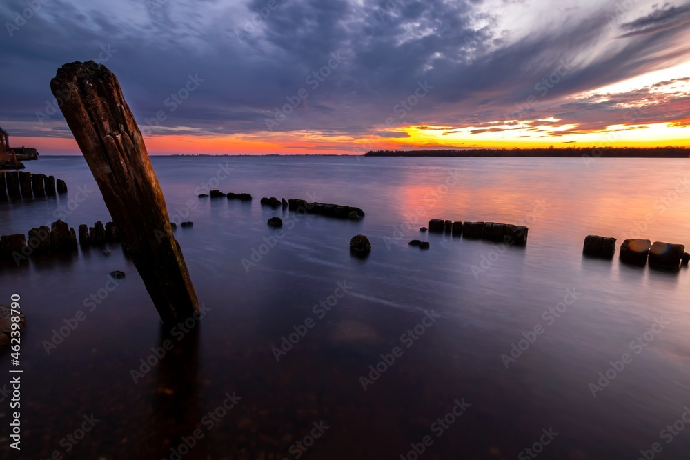 Beautiful sunset in the sky with clouds over the river. Lines of old wooden supports from the pier in the water in the foreground.