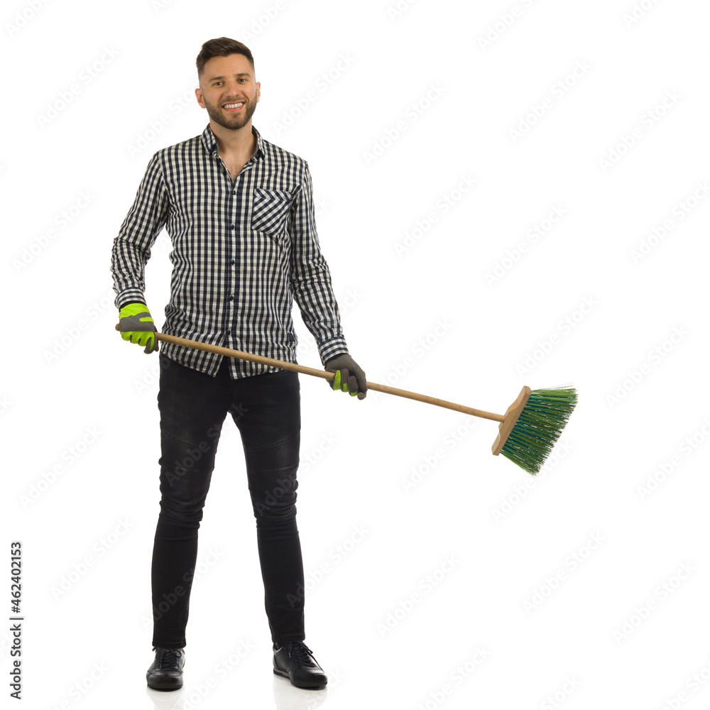 Handsome young smiling man is standing with broom. Full length, isolated.