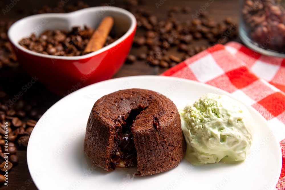 A portion of chocolate fondant with a scoop of pistachio ice cream on a plate. In the background is a heart-shaped cup filled with coffee beans and a cinnamon stick. To the right is a checkered napkin