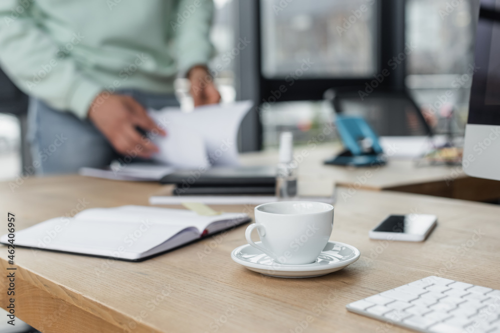 Fototapeta premium Cropped view of cup of coffee near computer and blurred businessman in office